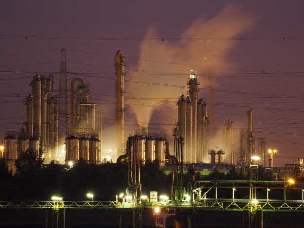 Ölraffinerie bei Nacht mit Rauchentwicklung, beleuchtet von umliegenden Lichtern, mit sichtbarem Himmel und Wasserzeichen.