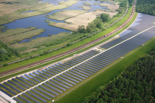 Luftaufnahme einer Solar-Farm mit Solar-Panelen, einem Zug auf einer Schiene, umgeben von Bäumen, Gras, Pflanzen und Wasser.
