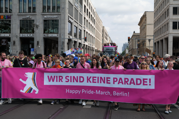 Eine Gruppe von Menschen marschiert eine Straße in Berlin entlang und hält eine pinkfarbene 'Happy Pride March'-Fahne hoch, mit Gebäuden, Laternen und Verkehrssignalen an der Straße unter einem bewölkten Himmel.