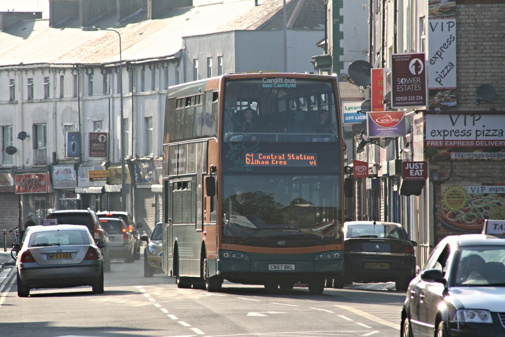 Eine Straße mit Autos und einem Bus vor Gebäuden mit Wänden, Fenstern, Tellern und Dächern, mit Plakaten und Bannern an den Wänden und einem Mast mit einer Straßenlaterne.