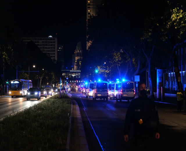 Eine nächtliche Straßen Szenerie mit Polizeiwagen und Bussen, die fahren, Menschen auf dem Bürgersteig gehen, Gras auf der linken Seite, Bäume und Gebäude im Hintergrund und Laternen die Straße beleuchten.