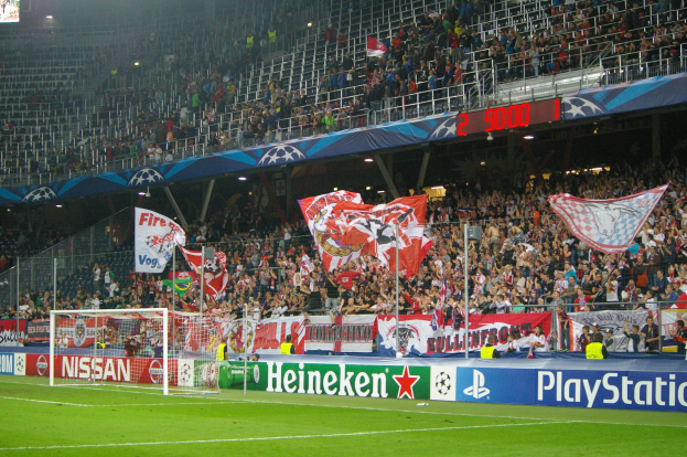 Ein Fußballspiel in einem hell erleuchteten Stadion mit einer großen Zuschauermenge, einem Torpfosten in der Mitte, Bannern drumherum und einem Bildschirm oben links.