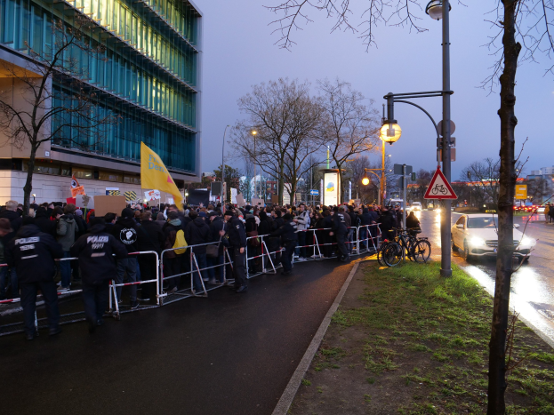 Eine große Gruppe von Menschen mit Schildern steht vor einem Gebäude mit Barrikaden und Bäumen, was auf eine Demonstration in Berlin hindeutet.