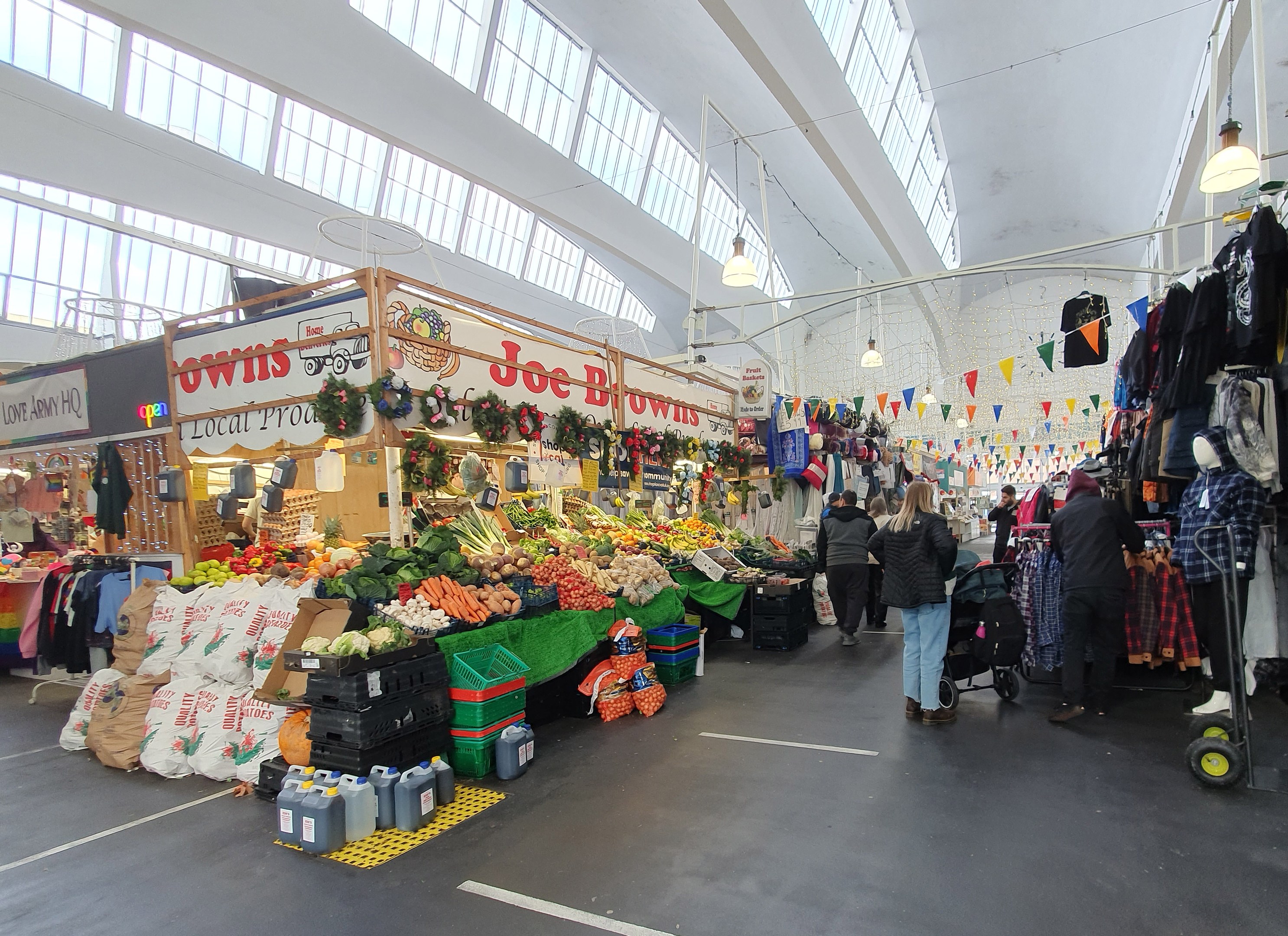 Innenmarkt mit Menschen, die an Ständen mit Obst, Gemüse und aufgehängten Kleidern unter Deckenlampen browsen, mit großen Glasfenstern im Hintergrund.