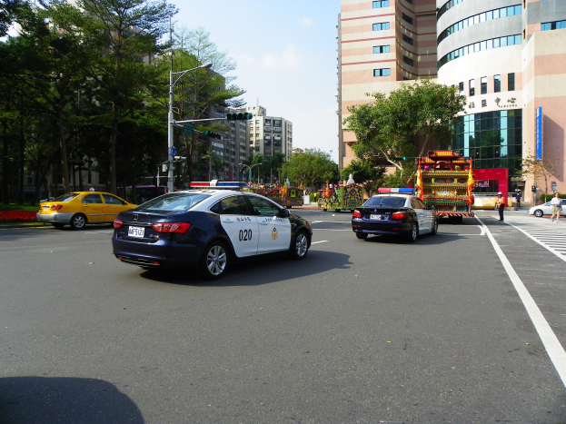 Ein Polizeiwagen fährt auf einer Straße mit hohen Gebäuden, Bäumen, Laternen, Verkehrszeichen und Menschen unter einem bewölkten Himmel.