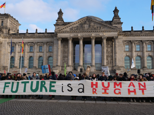 Eine Gruppe von Menschen hält ein Transparent mit der Aufschrift 'Zukunft ist ein Menschenrecht' vor dem Reichstaggebäude in Berlin, Deutschland, mit sichtbaren Fenstern, Säulen und Bögen des Gebäudes und nahen Fahnen mit Stangen unter einem bewölkten Himmel.