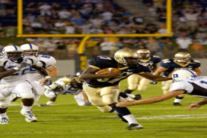 Ein Fußballspieler in einem Sportoutfit und Helm läuft mit dem Ball, während er von anderen Spielern umgeben ist, mit unscharfen Hintergrundelementen und Text am unteren Rand.