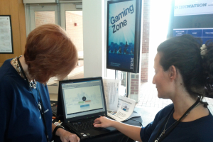 Eine Frau arbeitet an einem Laptop an einem Schreibtisch mit Papieren und einem Glasfenster daneben, mit einer Tür und einer Backsteinwand im Hintergrund.