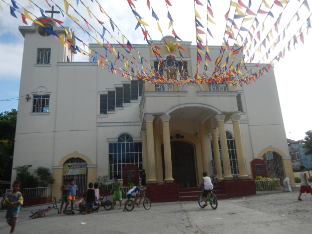 Eine Gruppe von Menschen, die Fahrräder vor einer weißen Kirche mit architektonischen Details, Pflanzen, einem Zaun, einer Fahne, Bäumen und einem bewölkten Himmel fahren, mit bunten Fahnen, die an einem Seil oben befestigt sind.