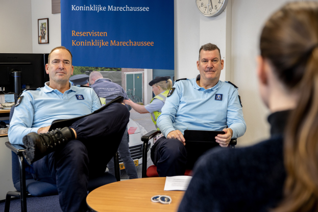 Zwei Polizeibeamte sitzen an einem Tisch mit einer Frau, Papieren und Gegenständen auf dem Tisch, einer Wandbanner und Uhr im Hintergrund, einem zweiten Tisch mit Monitor, Tastatur, Maus und Gegenständen links daneben und Fotorahmen an der Wand.