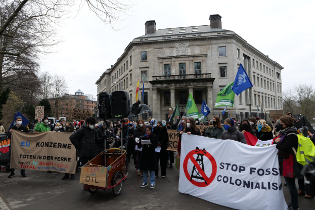 Große Gruppe von Menschen marschiert bei einer Demonstration gegen fossile Brennstoffe, trägt Schilder und Fahnen, mit einem Fahrzeug im Vordergrund, Bäumen und Gebäuden im Hintergrund unter einem klaren blauen Himmel.