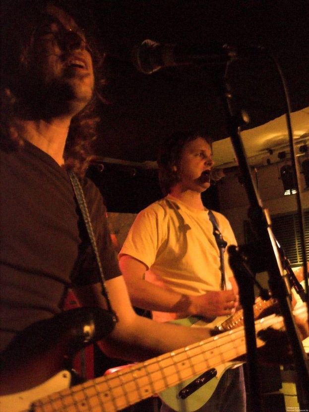 Ein Musikkonzert mit zwei Sängern: einer in einem weißen Hemd, das eine grüne Gitarre hält, und ein anderer in einem schwarzen T-Shirt mit Brille, der eine schwarze Gitarre hält, mit Bühnenbeleuchtung und Instrumenten im Hintergrund.