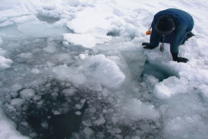 Eine Person kniet rechts im Bild auf den Schnee und schaut auf das Eis in der Mitte des Wassers.