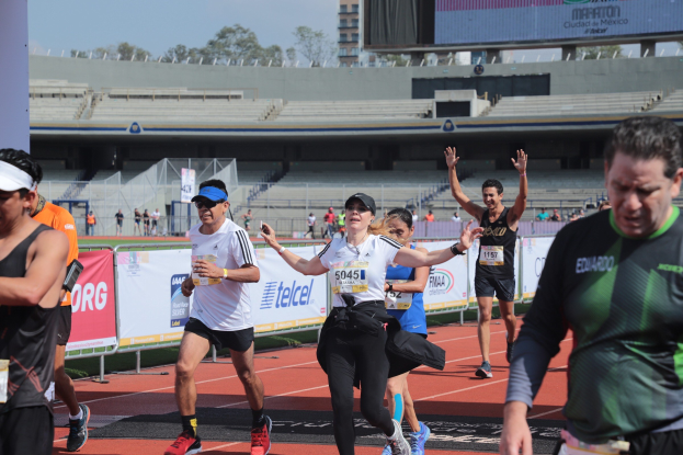 Gruppe von Menschen, die auf einer Stadionbahn laufen, mit Bannern, einem Bildschirm, einem Gebäude, Bäumen und einem bewölkten Himmel.