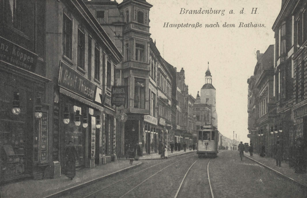 Ein altes Schwarz-Weiß-Foto einer Straßenkreuzung in Brandenburg, Deutschland, mit einer Tram auf den Schienen, Menschen auf dem Gehweg, Gebäuden mit Fenstern, Schildern und Straßenlaternen unter einem bewölkten Himmel.