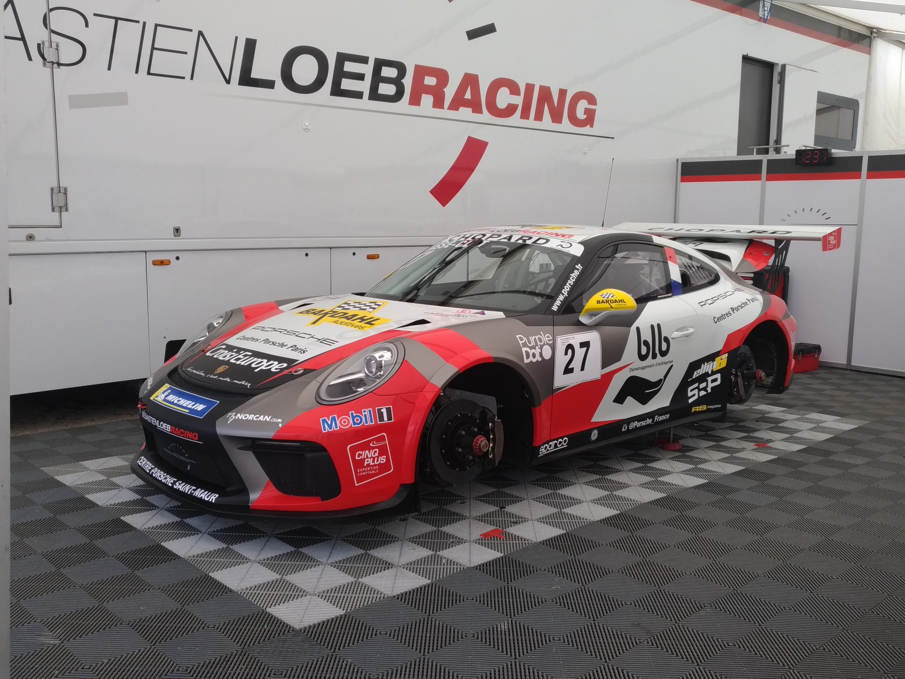 Ein Porsche 911 GT3 RSR in einer Garage, mit einer Wand im Hintergrund, die Text und eine Tür zeigt.