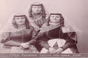Schwarzes und weißes Foto von drei Frauen in traditionellen russischen Hochzeitskleidern mit farbenfrohen Stickereien und Perlen, die vor einer weißen Wand stehen.