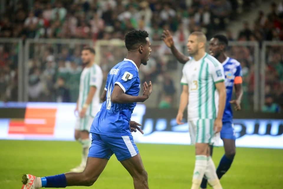 Ein Fussballspieler in einem blauen und weissen Jersey feiert mit erhobenen Armen, umgeben von einer Gruppe von Menschen in weissen und grünen Jerseys, mit einem unscharfen Hintergrund, der ein weisses und blaues Brett und ein Geländer zeigt.