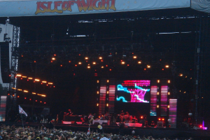 Eine Bühne mit Lichtern, einem Bildschirm und Musikern, die Instrumente spielen, mit einem Banner oben, einer Menge unten und Fahnen mit Stangen und Ballons drumherum.