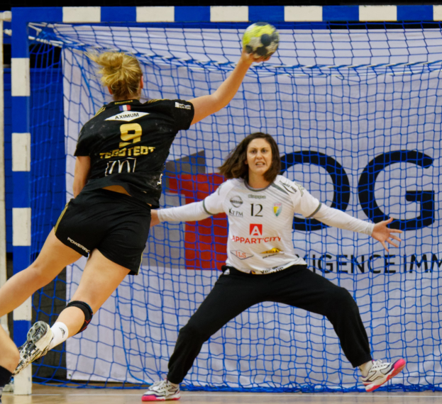 Zwei Frauen beim Handballspielen in der Nähe eines Tores, eine hält den Ball, mit Netz und einer Tafel im Hintergrund.