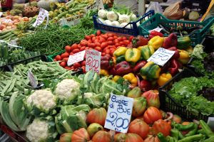 Ein belebter Bauernmarkt mit verschiedenen Gemüsen wie Tomaten, Paprika, Blumenkohl, grüne Bohnen in Körben, Preisetiketten sichtbar und einige Menschen anwesend.