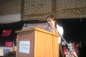 Ein Mädchen in einem weißen Hemd steht neben einem Rednerpult in einem Konferenzraum, rechts daneben ein Mikrofonständer, ein weißes Banner an einer Wand im Hintergrund und mehrere Taschen an einem Faden rechts.