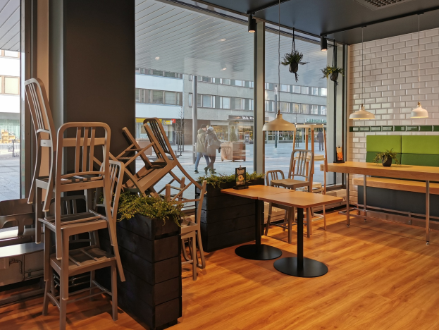 Ein Restaurant-Innenraum mit hölzernen Tischen und Stühlen in der Nähe eines großen Fensters, das eine Straßenszene in Berlin mit Passanten, Gebäuden und Straßenelementen zeigt.