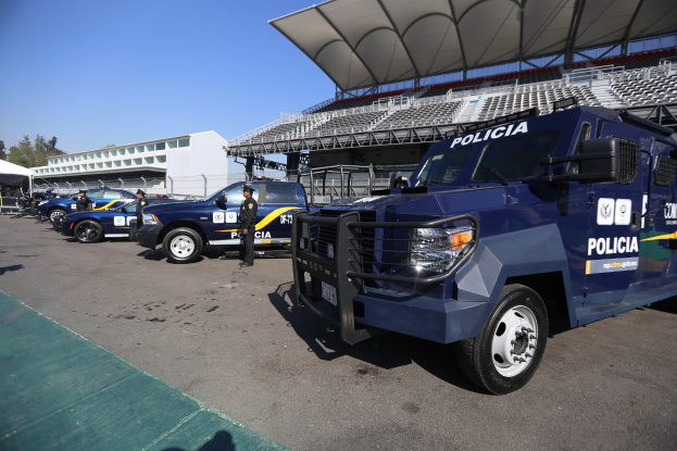 Eine Reihe von Polizeiwagen vor einem Stadion geparkt, mit Menschen auf der Straße im Vordergrund und Gebäuden, Bäumen und einem klaren blauen Himmel im Hintergrund.