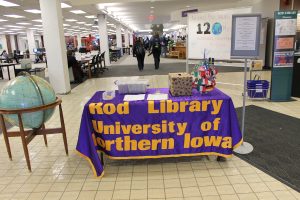 Eine Ausstellung mit einem Tisch, auf dem verschiedene Landesflaggen, eine Box, Papiere und ein Banner liegen, eine Kugel links daneben, ein Anschlagbrett rechts mit ein paar Leuten in der Nähe und mehreren Tischen und Stühlen links.