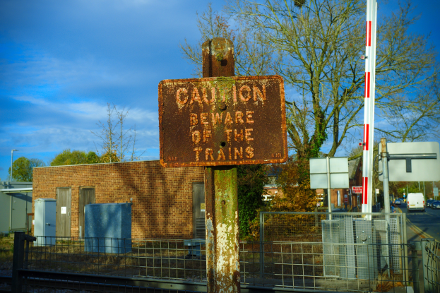 Warnschild auf einer Bahnschiene umgeben von einem Metallzaun, Bäumen, Strommästen, einem Gebäude mit Fenstern, Containern, Fahrzeugen auf einer Straße und einem bewölktem Himmel.