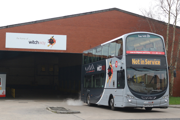 Doppeldeckerbus mit Rauchwolke an einem Gebäude vorbeifahrend, mit Gras, Bäumen und einem klaren Himmel im Hintergrund; sichtbares Kfz-Kennzeichen am Bus.