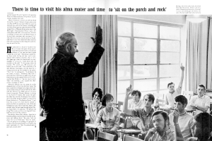 Eine Person mit Brille steht in einem Raum mit sitzenden Personen, Vorhängen und Fenstern.