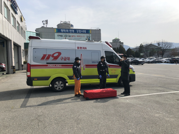 Rettungswagen auf einem Parkplatz neben einem Geb├Ąude mit drei Personen auf der Stra├če davor geparkt, glasfensteriges Geb├Ąude links, andere Fahrzeuge, B├Ąume, Berge und Himmel im Hintergrund.