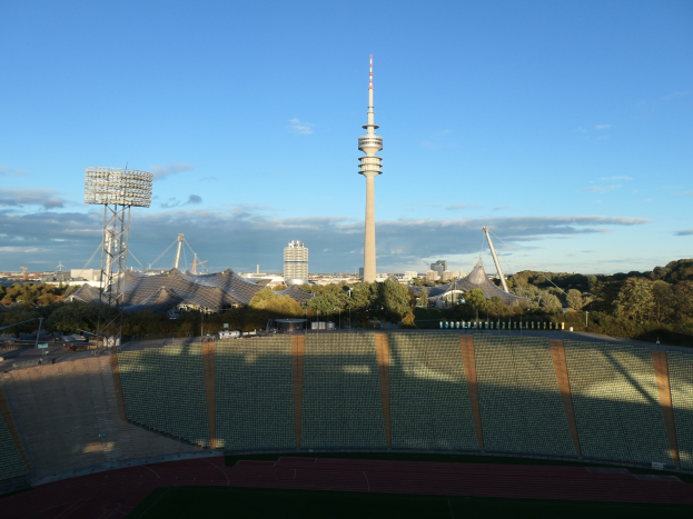 Olympiastadion in Berlin, Deutschland, mit dem Fernsehturm (Fernsehturm) im Hintergrund, umgeben von Bäumen, Gebäuden und Lichtern unter einem bewölkten Himmel.