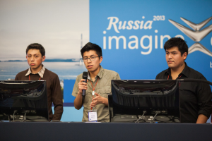Drei Jungen stehen neben einem Schreibtisch mit zwei Computersystemen, einer spricht in ein Mikrofon und trägt eine ID-Karte mit roter Markierung, vor einer Wand mit einer Werbetafel, auf der 'Russland Imagine 2013' steht.