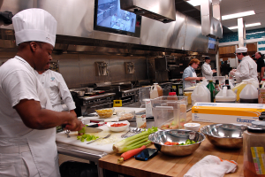 Eine Gruppe von Köchen in einer Küche, die auf einem Schneidbrett Essen zubereiten, mit einem Koch im Vordergrund in weißer Kleidung und Mütze, der ein Messer hält und von verschiedenen Utensilien, Schüsseln, Gemüse, einem Handy, Flaschen und anderen Gegenständen umgeben ist, mit einem Fernseher und Deckenlampen im Hintergrund.