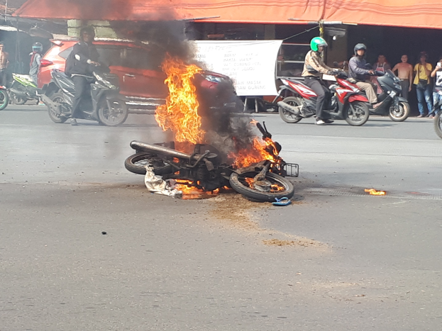 Motorrad in Flammen an der Straße mit Menschen, einem Auto, einem Van, einer Fahne und einem Zelt im Hintergrund.