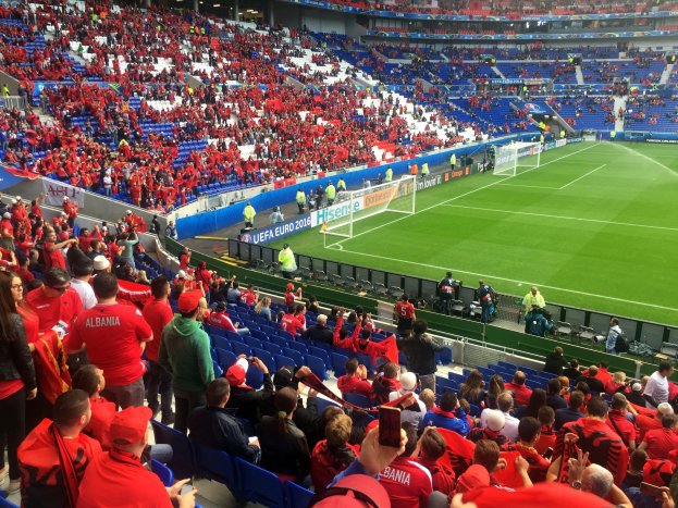 Eine große Menschenmenge in einem Stadion schaut einem Füßballspiel zu, mit Sitzplätzen und Stehplätzen, Bannern, einem Torpfosten und Deckenleuchten.