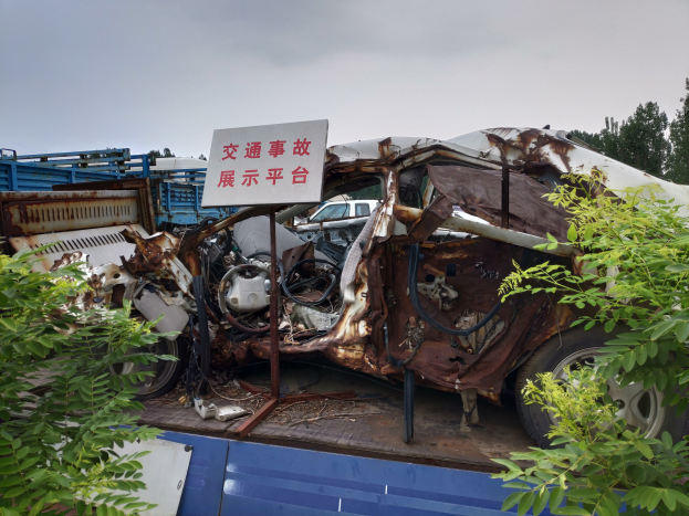 Ein verrostetes und verbeultes Auto auf einem Truck-Bett gesichert, flankiert von Grün, mit Bäumen und einem klaren blauen Himmel im Hintergrund.