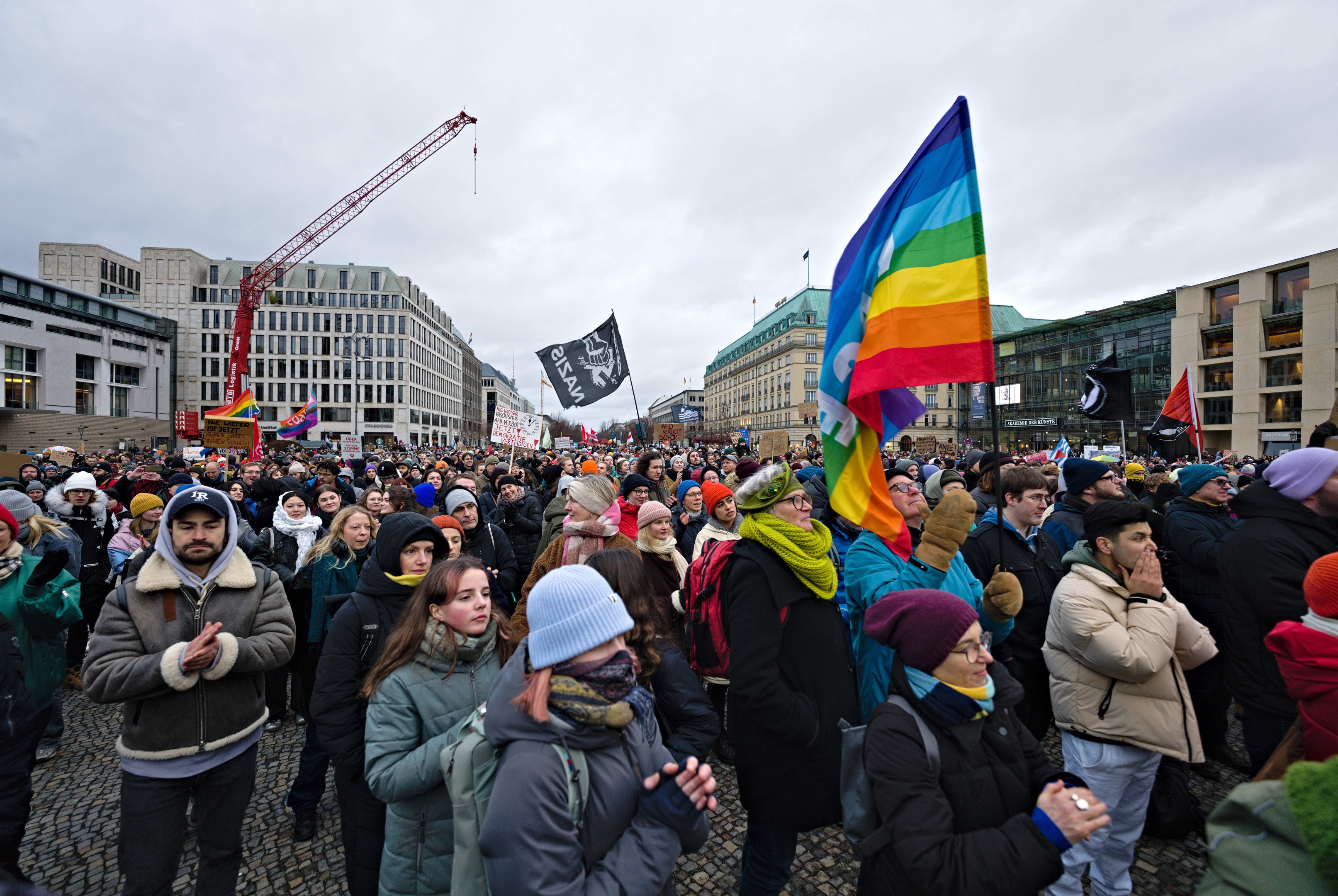 Große Gruppe von Menschen bei einer LGBTQ+-Rechtsdemo in Berlin mit Fahnen und Plakaten, im Hintergrund Gebäude, ein Kran und ein bewölkter Himmel.