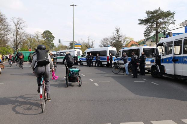 Gruppe von Menschen auf Fahrrädern auf einer Straße mit Polizeiwagen, anderen Fahrzeugen, Bäumen, Laternen, Verkehrsschildern, Schildern, Gebäuden und einem bewölkten Himmel.