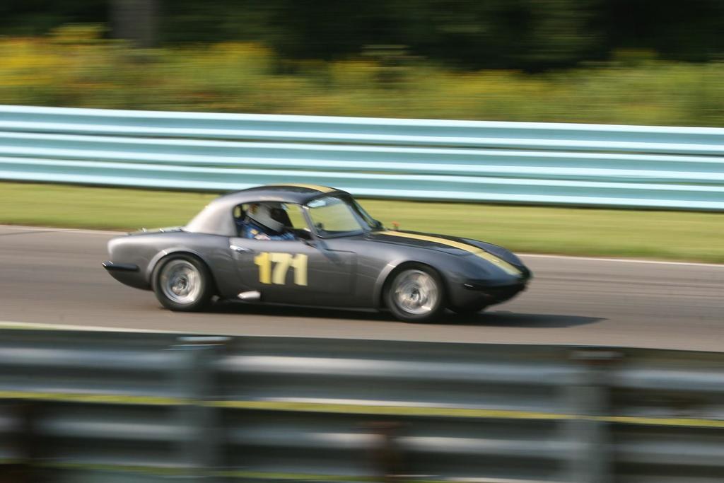 Ein Auto mit einem Helm tragenden Fahrer fährt eine von Barrieren gesäumte Straße entlang, mit Bäumen im Hintergrund.