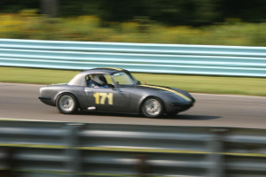 Ein Auto mit einem Helm tragenden Fahrer fährt eine von Barrieren gesäumte Straße entlang, mit Bäumen im Hintergrund.