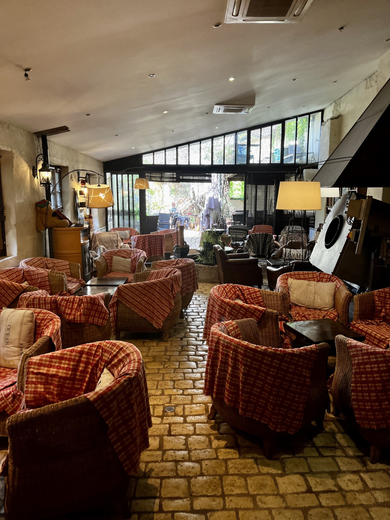 Ein geräumiger Eingangsbereich mit zahlreichen Stühlen, Tischen, gepolsterten Sofas, an der Wand montierten Lampen, einer Deckenklimaanlage und Glasfenstern an der Rückseite, identifiziert als der Empfang des Hotel de Ville.