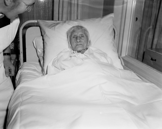 Alte Frau im Krankenhausbett liegend mit einer Krankenschwester daneben, Bett mit einem weißen Laken und Kissen bedeckt, Fenster mit Vorhängen im Hintergrund.