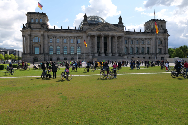 Eine Gruppe von Menschen, die vor dem Reichstaggebäude in Berlin, Deutschland, Fahrrad fährt, mit sichtbaren Fenstern, Säulen und Fahnenmasten des Gebäudes, umgeben von Grün unter einem bewölkten Himmel.