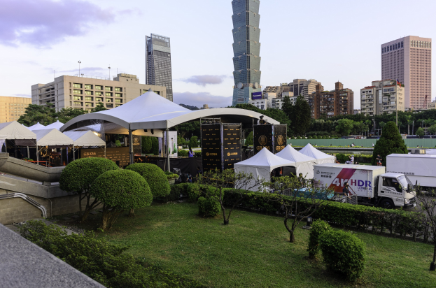 Eine Gruppe von Zelten in einem Park mit Grünflächen, Menschen, Fahrzeugen, Gebäuden und einem klaren Himmel, wahrscheinlich auf dem Hong Kong International Film Festival.