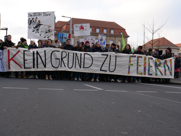 Demonstranten mit einer Transparent haltend, auf dem 'Kein Grund zu Feiern' steht, stehen auf einer Straße mit Straßeninfrastruktur und Gebäuden im Hintergrund bei klarem Himmel.