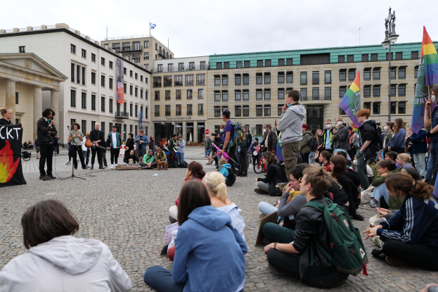 Menschen sitzen auf dem Boden vor einer Menge mit Fahnen und Spruchbändern bei einer anti-schwulen Demonstration in Berlin, mit Mikrofonständer, Statue auf einem Sockel und Gebäuden im Hintergrund.