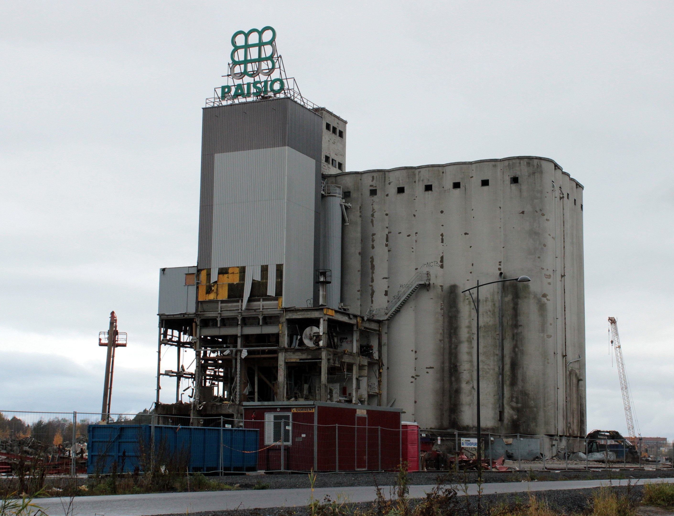 Großer Zementbau neben einer Straße, umgeben von Pflanzen, einem Zaun, Pfählen, Lampen und anderen Gegenständen, mit Bäumen und einem klaren blauen Himmel im Hintergrund.